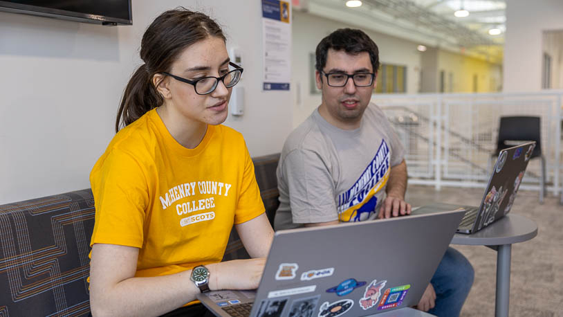 Two MCC students are working on laptops in the open, brightly lit Foglia CATI building.