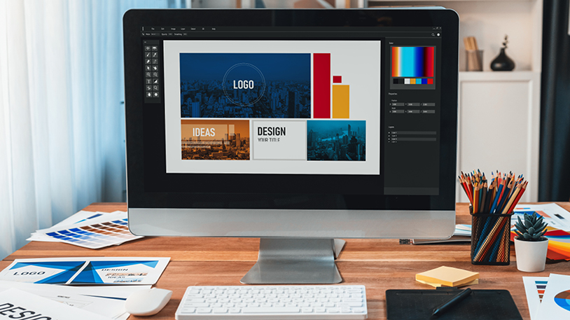 A computer monitor on a desk displays graphic design software with a project open featuring a city-themed layout, while the workspace is filled with design tools, including colored pencils and printed design samples.