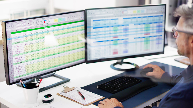 A person working at a desk with dual monitors displaying complex financial spreadsheets, with a clipboard, pen, and office supplies nearby.