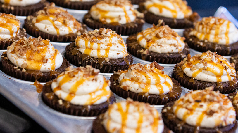 A close-up view of a tray of freshly baked chocolate cupcakes topped with frosting, caramel drizzle, and chopped nuts.