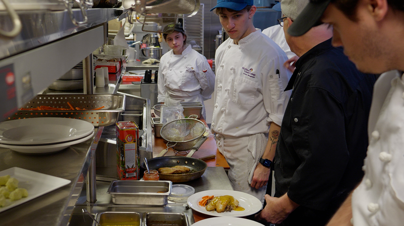 Culinary students and an instructor in McHenry County College uniforms working in a professional kitchen, with a plated dish in the foreground.