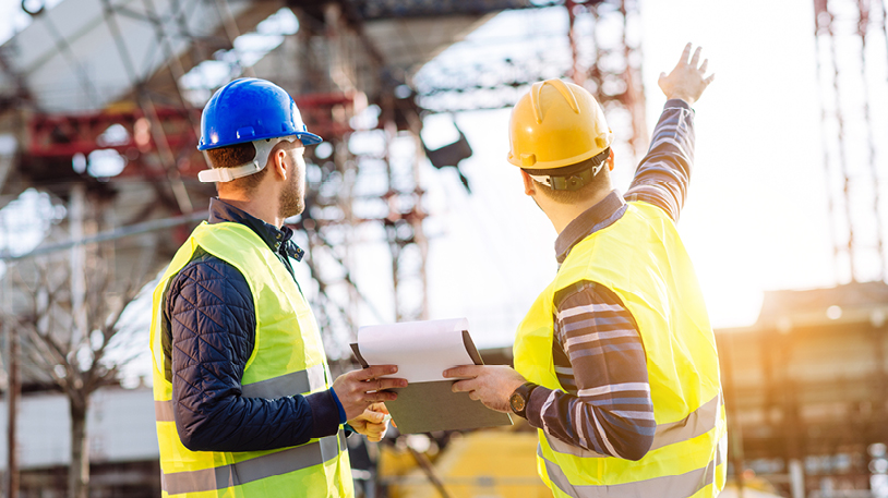 Two construction professionals in hard hats and high-visibility vests reviewing documents on a clipboard at an active building site.
