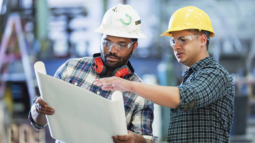 two males wearing hard hats look at blue prints
