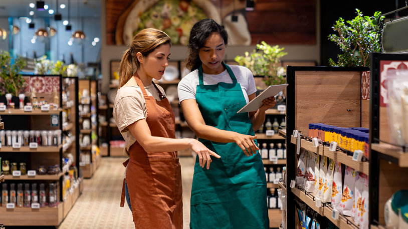 Two retail employees wearing aprons discussing inventory while looking at a tablet in a store aisle.
