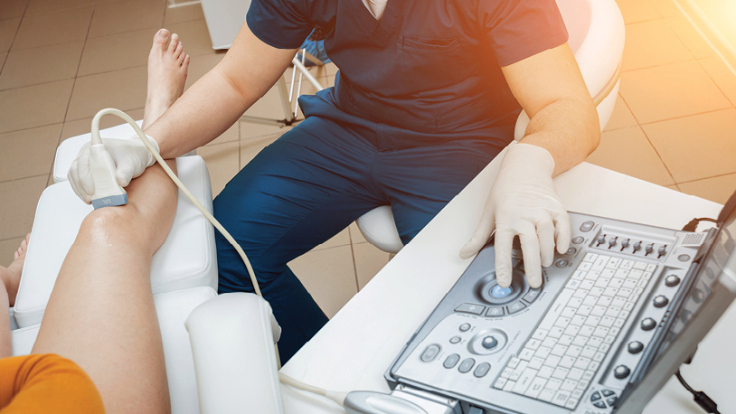 A medical professional wearing gloves is performing an ultrasound scan on a patient's lower leg using a handheld probe and an ultrasound machine.