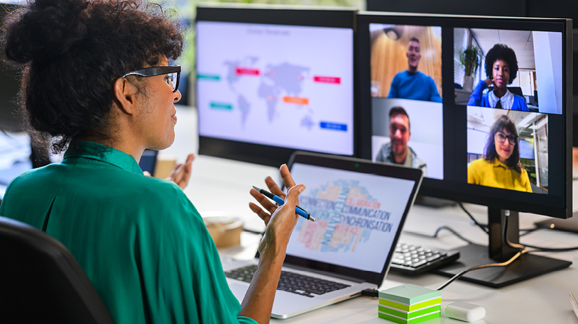 A professional participating in a remote video conference while working on a laptop and multiple monitors in an office.