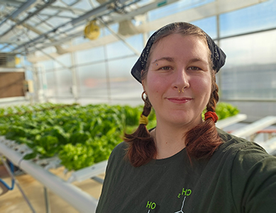 MCC horticulture student Jyinxs Crisp is wearing a dark green shirt and bandana, standing inside MCC's greenhouse with rows of hydroponically grown leafy green plants.