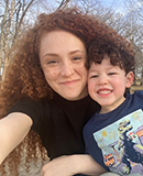 MCC alumna Katherine Sanders smiles as she hugs a cheerful young boy wearing a dinosaur-print sweatshirt outdoors on a sunny day.