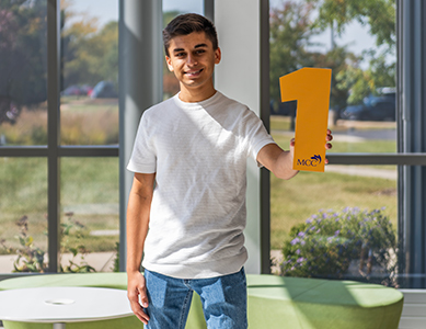 MCC student Humza Bangash stands in the lobby of the Liebman Science Center holding a number 1. 