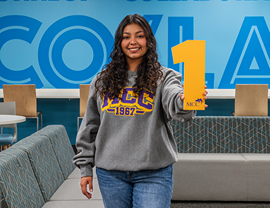 MCC student Yadira Placencia, wearing a gray MCC 1967 sweatshirt, holds a yellow number 1 cutout in Co/Lab B.